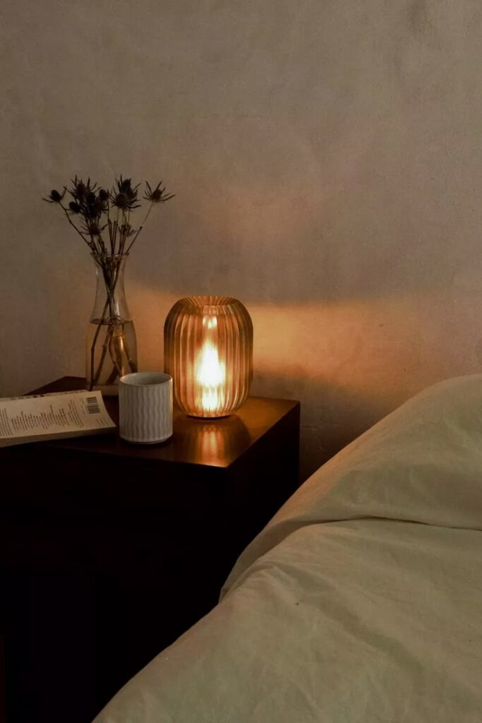 Cozy bedroom scene with a glowing lamp, vase with flowers, mug, and book on a wooden nightstand, beside a soft bed.