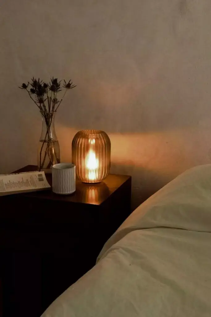 Cozy bedroom scene with a glowing lamp, vase with flowers, mug, and book on a wooden nightstand, beside a soft bed.