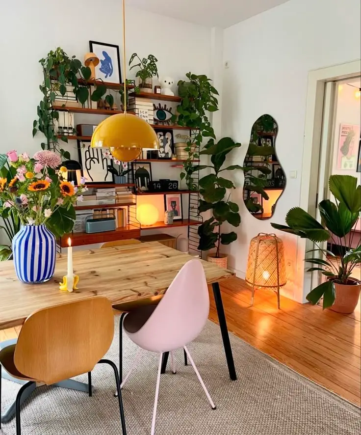 Stylish dining area with colorful decor, wooden table, mixed chairs, vibrant plants, and a unique mirror on a hardwood floor.