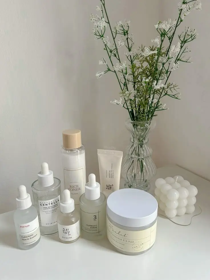 Skincare products with minimalist packaging on a white surface, accompanied by a vase of flowers and a decorative candle.