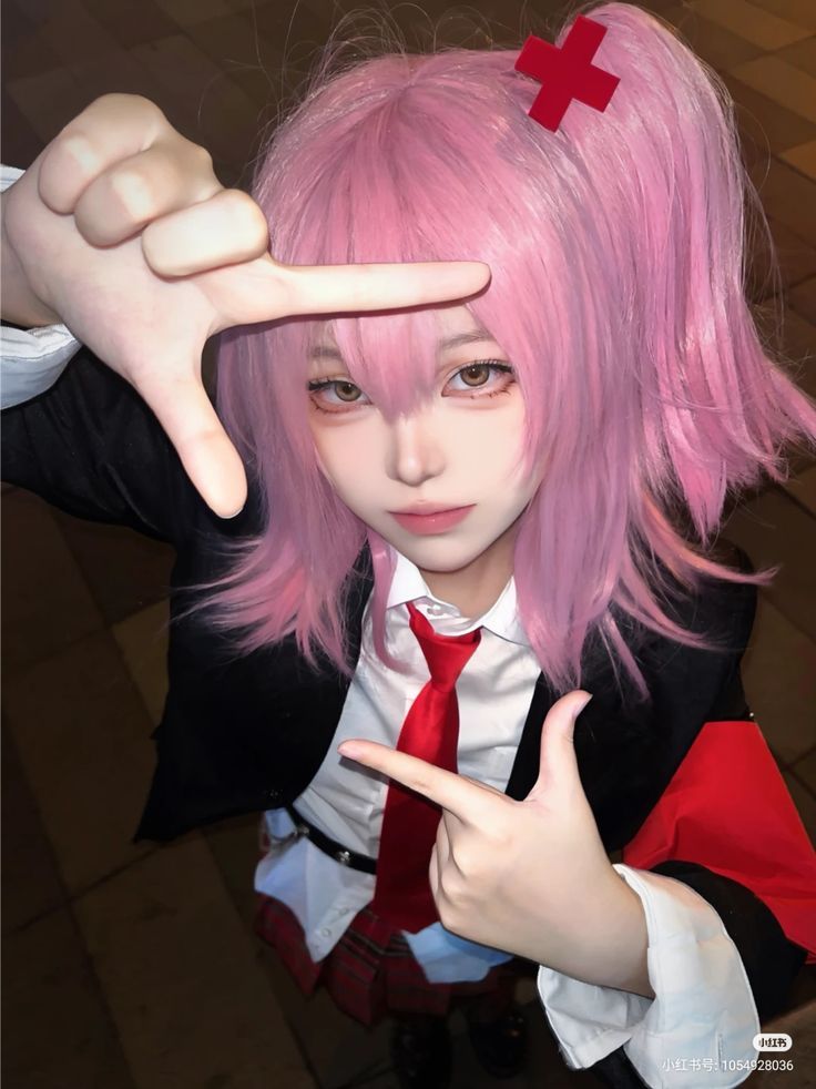 Person in pink wig and cosplay outfit making a finger frame gesture. Red Cross hairpin and plaid skirt complete the look.