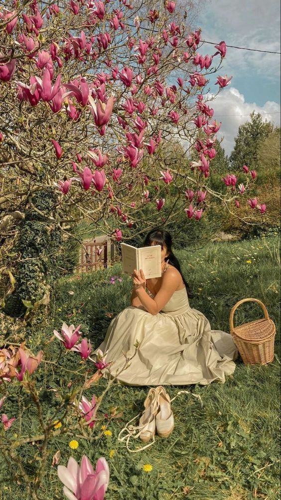 Woman reading under blooming magnolia tree with a picnic basket nearby, creating a peaceful springtime scene.