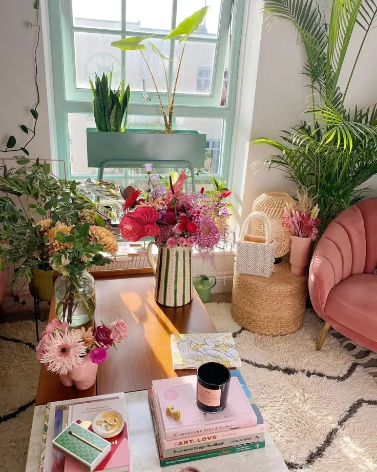 Cozy room with vibrant flowers, lush plants, a pink chair, and books on a wooden table. Bright, inviting, and stylish interior.