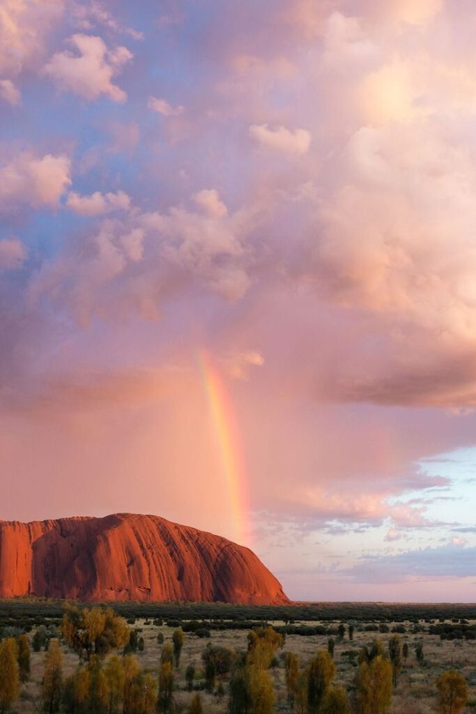 Scenic view of Uluru at sunset with a vibrant rainbow in a colorful sky, surrounded by natural Australian landscape.