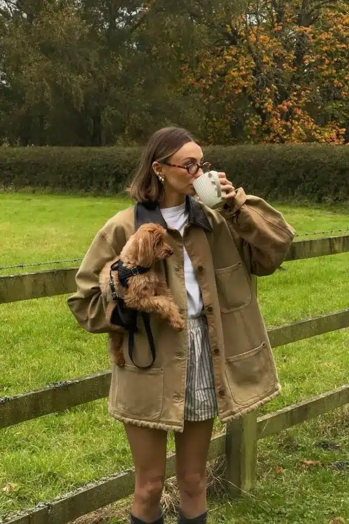Woman enjoys coffee outdoors, holding small dog. Dressed in beige jacket and stylish glasses. Autumn vibes in a green park.