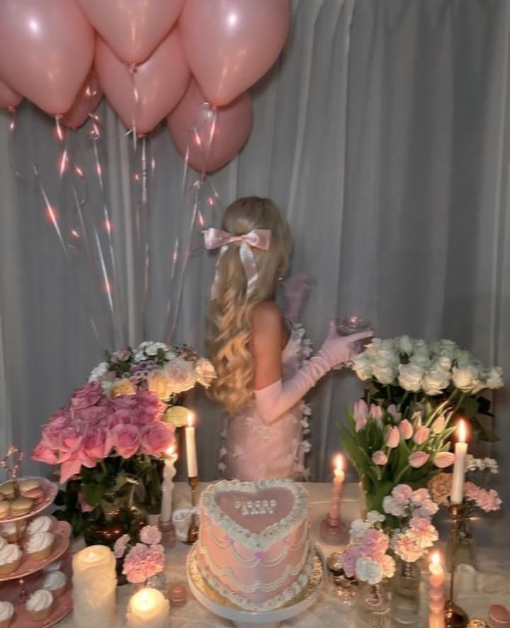 Woman in elegant dress at a pink-themed celebration with balloons, flowers, a heart-shaped cake, candles, and treats.