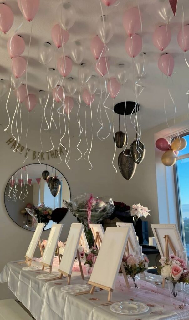 Birthday party setup with canvases, pink balloons, flowers, and a Happy Birthday banner in a decorated room for painting activity.