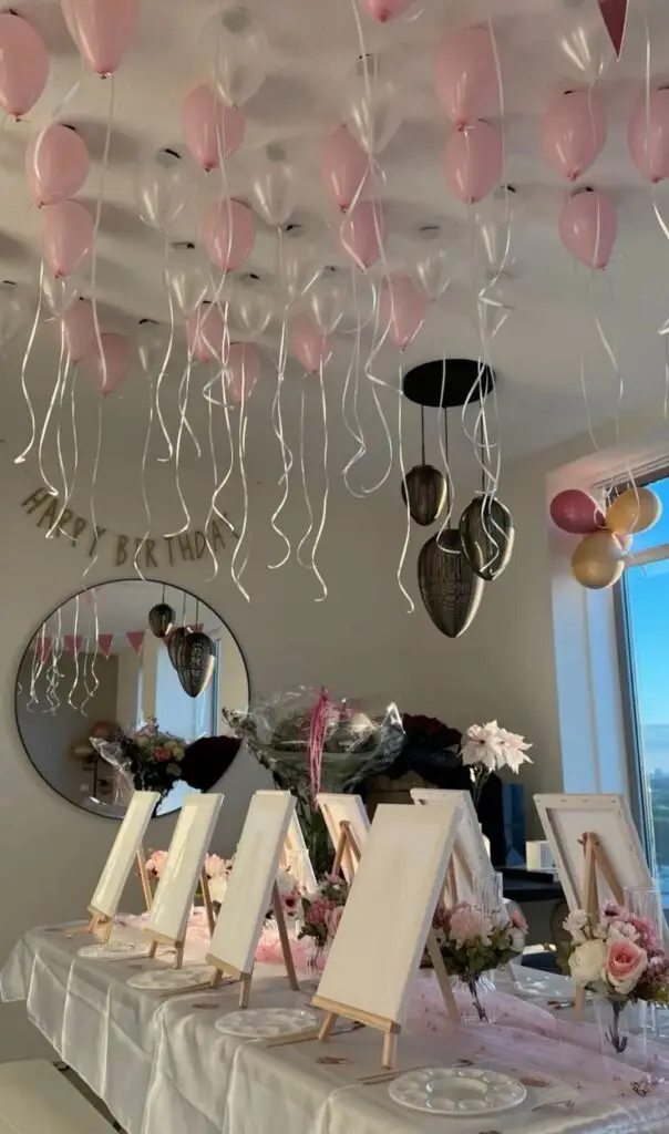 Birthday party setup with canvases, pink balloons, flowers, and a Happy Birthday banner in a decorated room for painting activity.