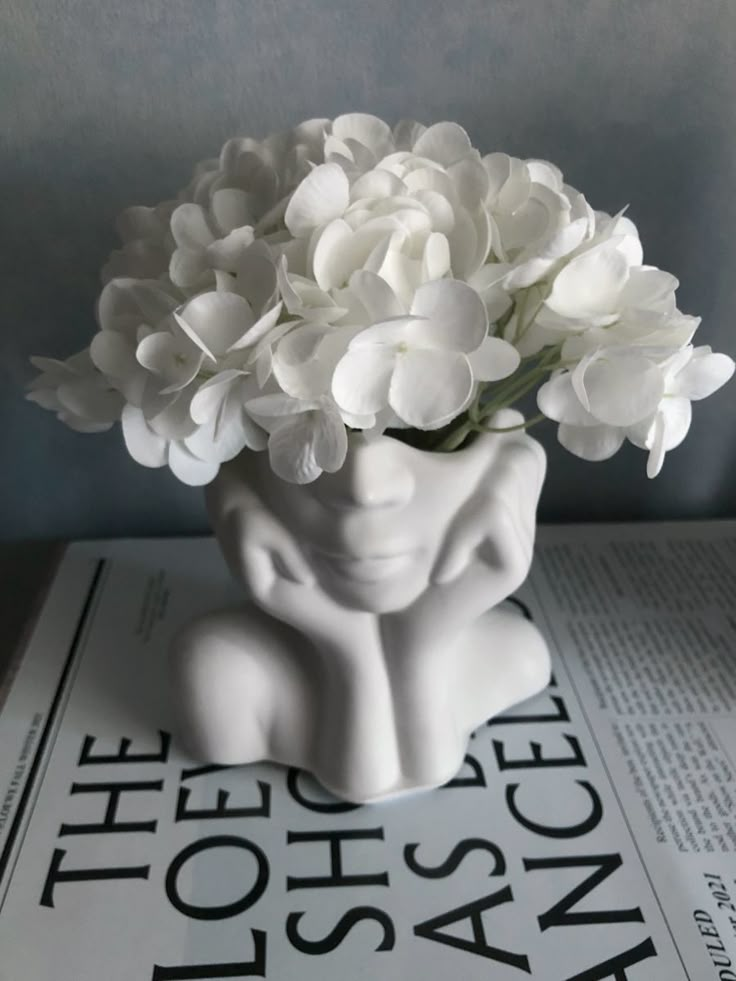 White hydrangeas in a sculpted face vase on a magazine, creating a modern and artistic decor piece.