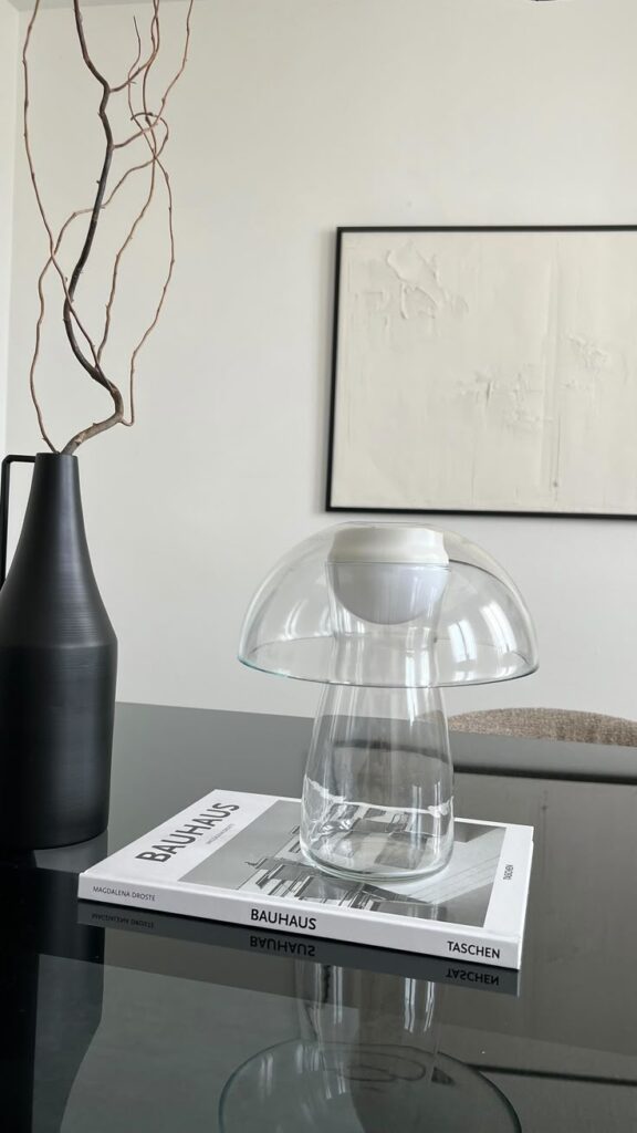 Modern glass lamp on a Bauhaus book beside a black vase with branches, minimalist decor on a black table with abstract wall art.