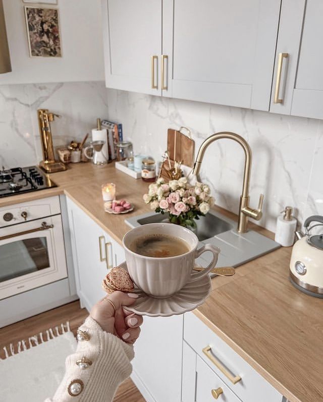 Cozy kitchen with coffee cup and macarons, elegant decor, chic white cabinets, wooden countertop, stylish gold faucet, and flowers.