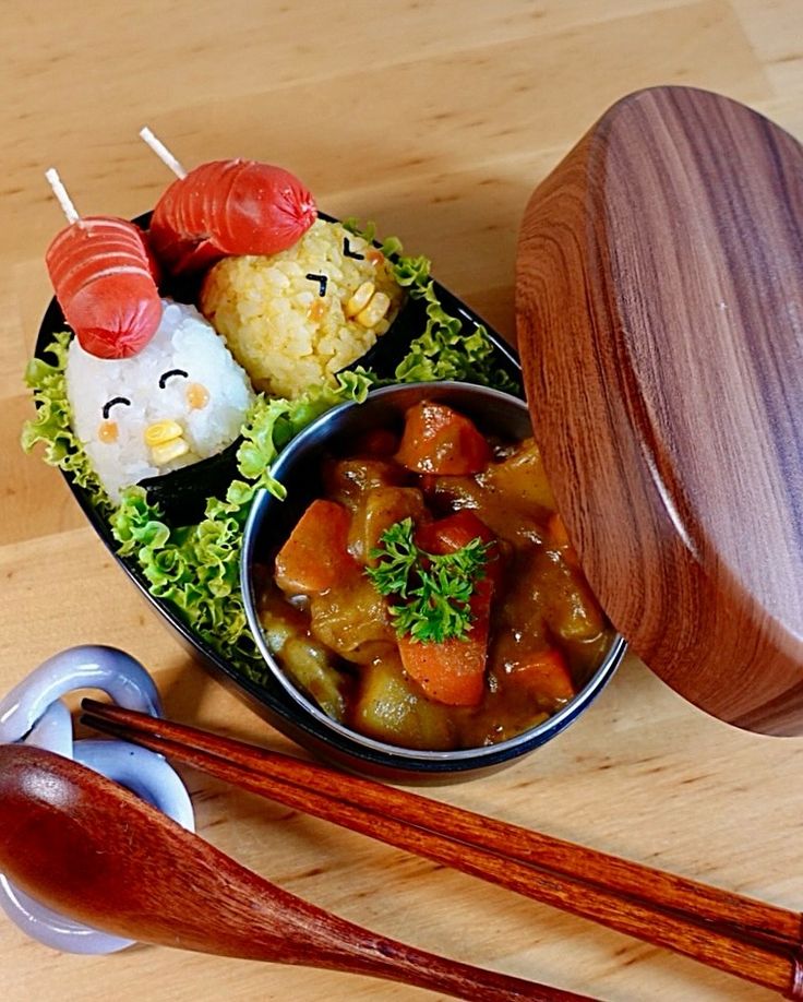 Creative bento box with cute rice ball birds and savory curry, garnished with fresh lettuce and herbs. Wooden lid and utensils included.