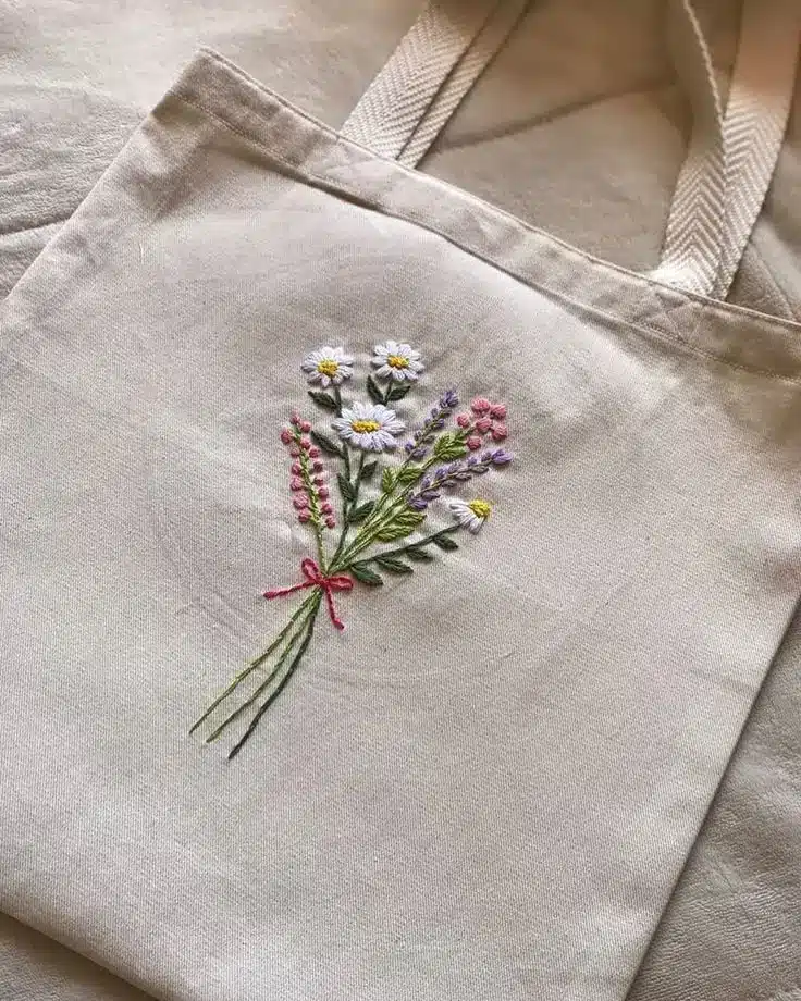 Tote bag with embroidered floral bouquet design, featuring daisies and lavender, on a beige fabric background. Eco-friendly fashion.