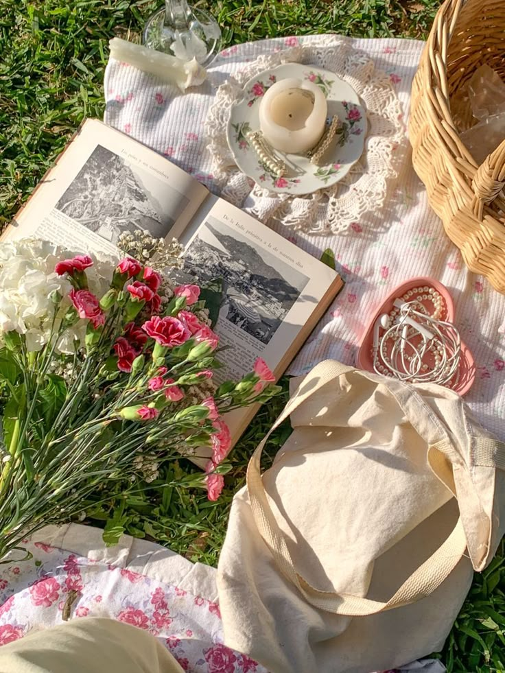 Picnic setup with open book, fresh flowers, candle, and basket on floral blanket in sunlight. Cozy outdoor reading and relaxation.