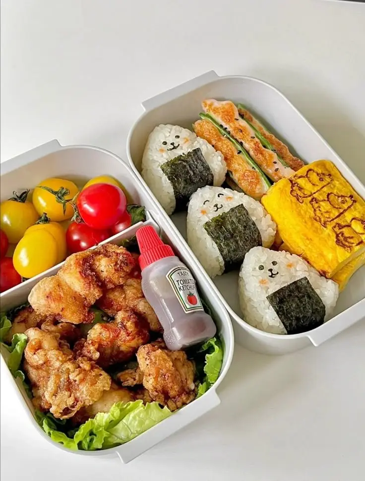 Cute bento box with rice balls, fried chicken, tomatoes, lettuce, and omelette. Adorable lunch presentation with ketchup bottle.