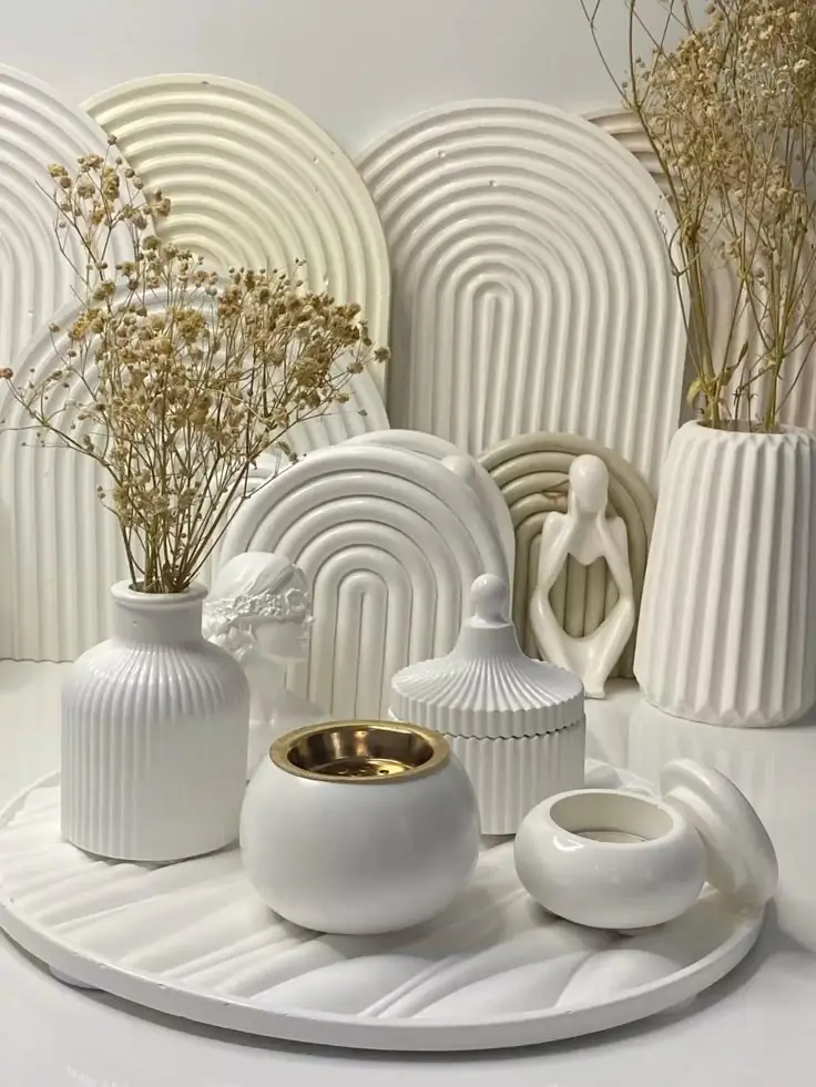 Modern ceramic decor with vases, sculpted figures, and dry flowers on a textured surface. Minimalist white and gold accents.