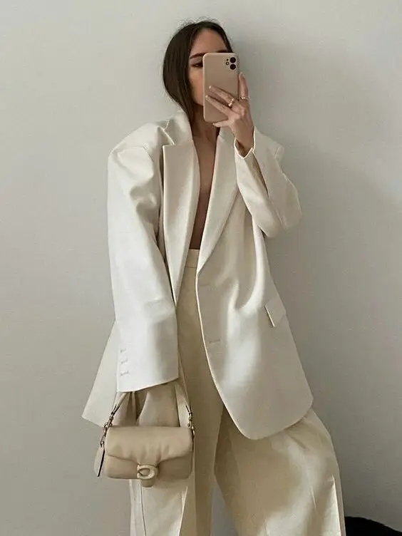 Stylish woman in oversized white suit takes a selfie, holding a beige handbag, showcasing chic minimalist fashion.
