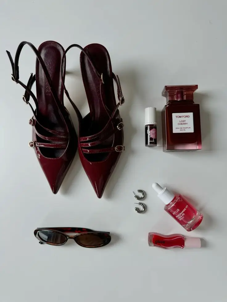 Stylish burgundy heels paired with Tom Ford perfume, lipstick, serums, sunglasses, and earrings on a flat lay background.