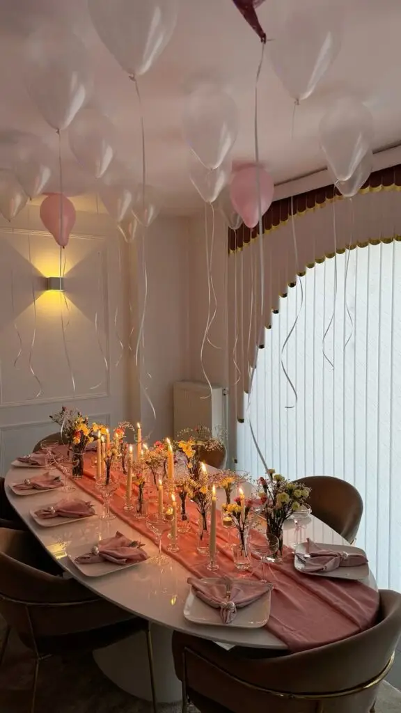 Elegant party table setting with pink decor, candles, and floral arrangements. White and pink balloons float above.