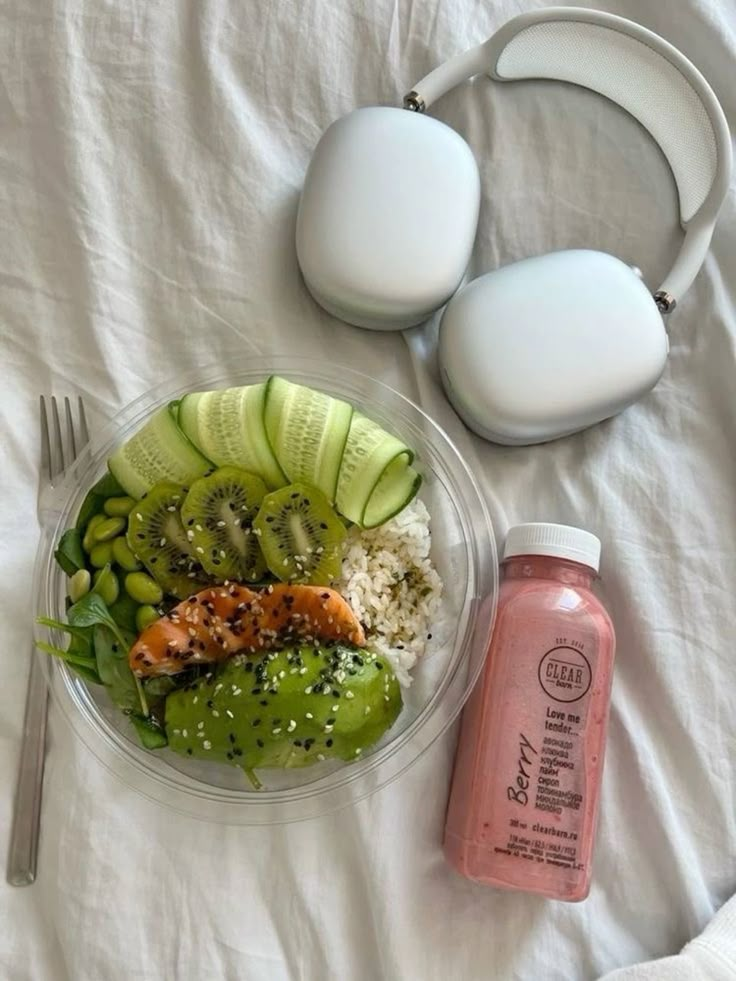 Healthy meal with rice, salmon, avocado, kiwi, cucumber, and edamame, alongside pink smoothie and headphones on a white sheet.