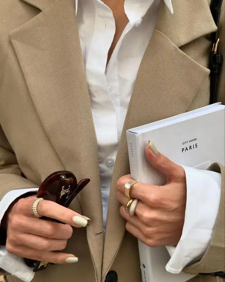 Stylish outfit with beige blazer, holding sunglasses and a Paris guidebook, showcasing elegant gold rings and chic manicure.