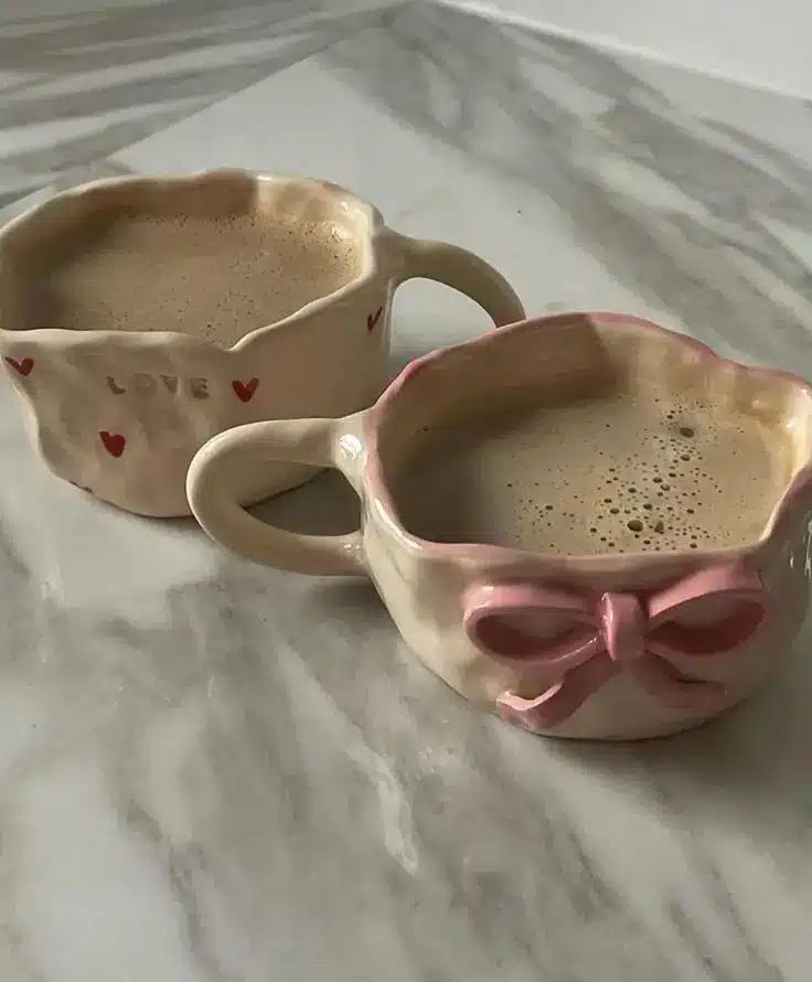Two whimsical mugs of coffee on a marble surface, one with a pink bow, the other with red hearts and LOVE text. Cozy morning vibes.