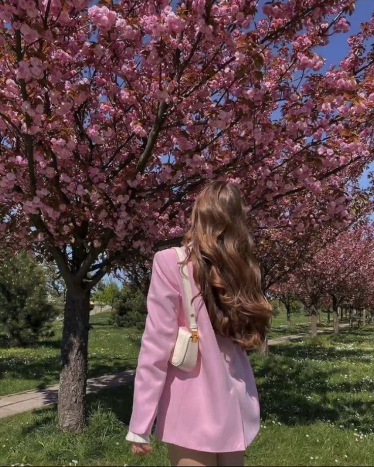 Woman in pink coat walking under blooming cherry blossom trees in sunny park, enjoying springtime beauty.