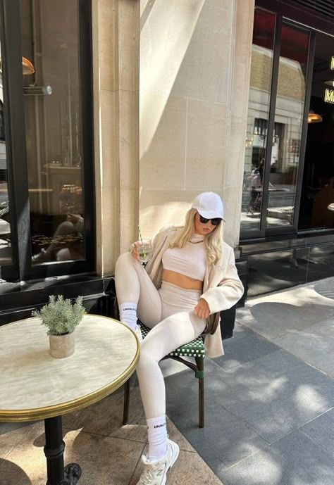 Trendy woman in white athleisure outfit, sitting at a cafe, wearing sunglasses and cap, enjoying a sunny day.