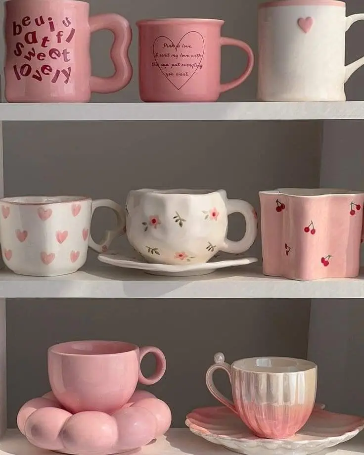 Collection of pink and white ceramic mugs and teacups with heart and floral designs on a shelf, perfect for cozy decor.