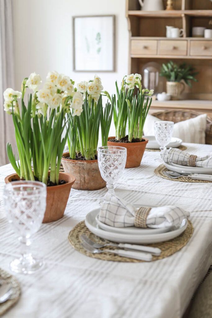 Elegant table setting with white flowers in terracotta pots, crystal glasses, and neatly arranged plates on a woven mat.