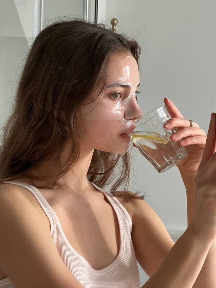 Woman wearing hydrating face mask, drinking lemon water. Skincare routine at home, self-care and wellness concept.