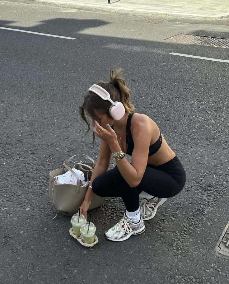 Woman in workout gear squatting on road, holding iced drinks, wearing headphones. Fitness and refreshment on a sunny day.