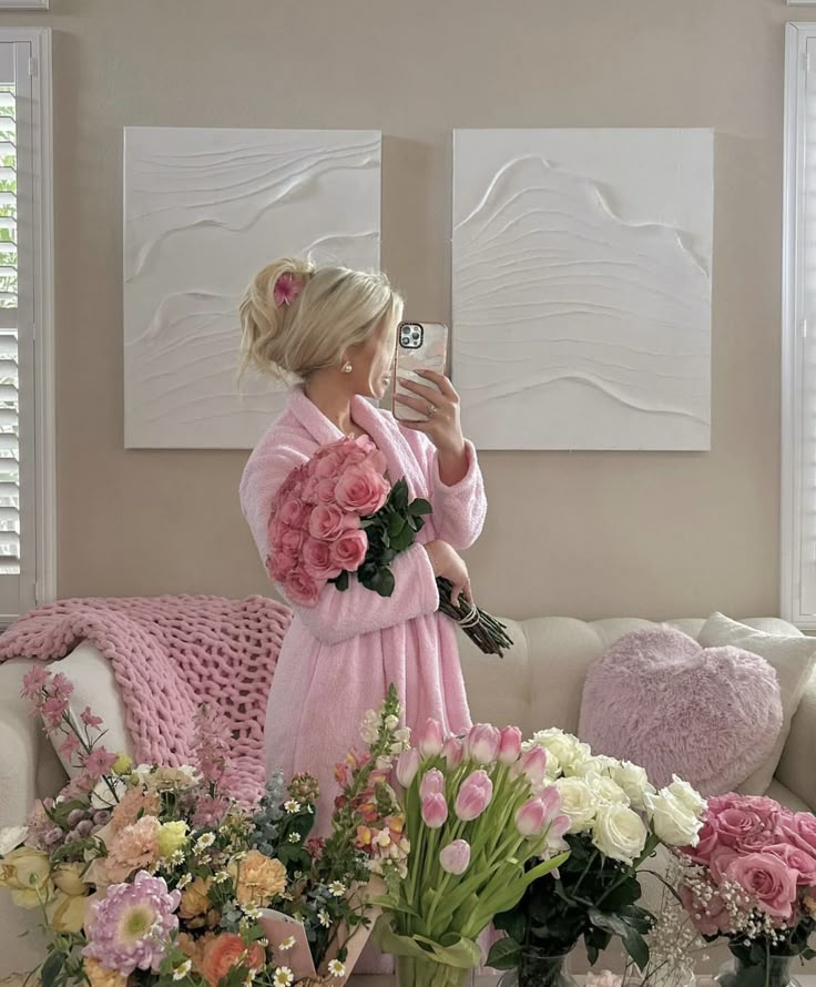 Person in pink robe holding roses in a cozy room with floral decor and abstract wall art, capturing a self-portrait with a phone.