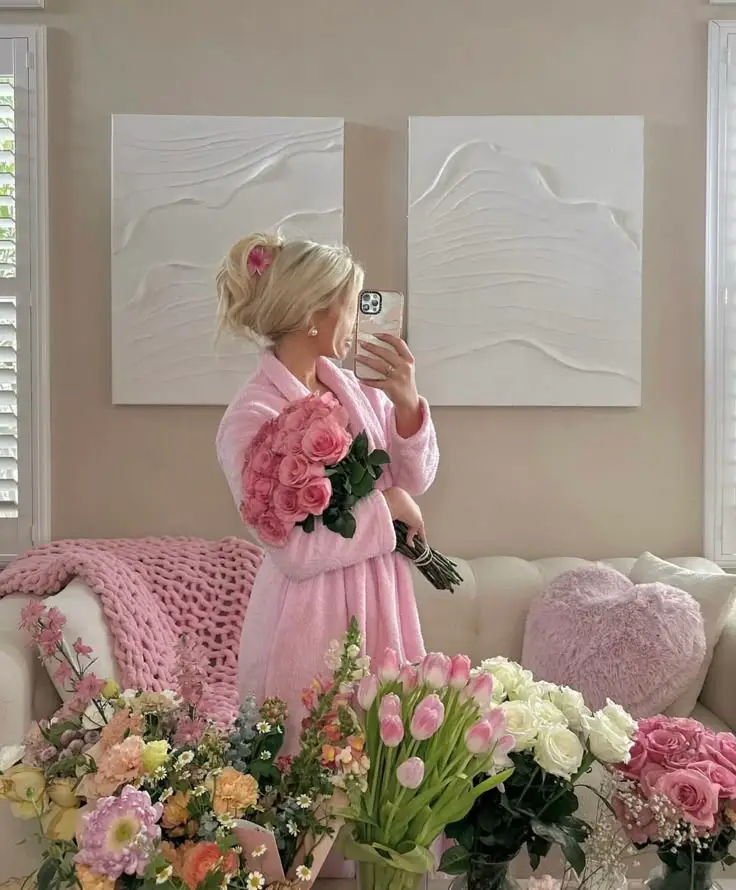 Person in pink robe holding roses in a cozy room with floral decor and abstract wall art, capturing a self-portrait with a phone.