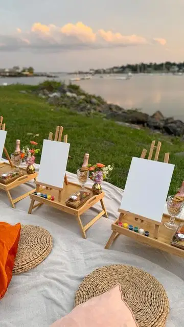 Outdoor painting setup by the serene lakeside with easels, paint, and champagne under a picturesque sunset sky. Perfect for artists.