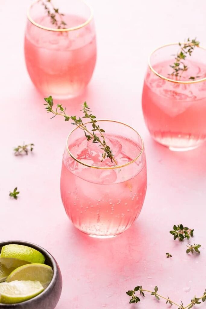 Refreshing pink cocktails with ice, garnished with thyme sprigs, on a pastel surface. Lime wedges in a bowl add a citrus touch.