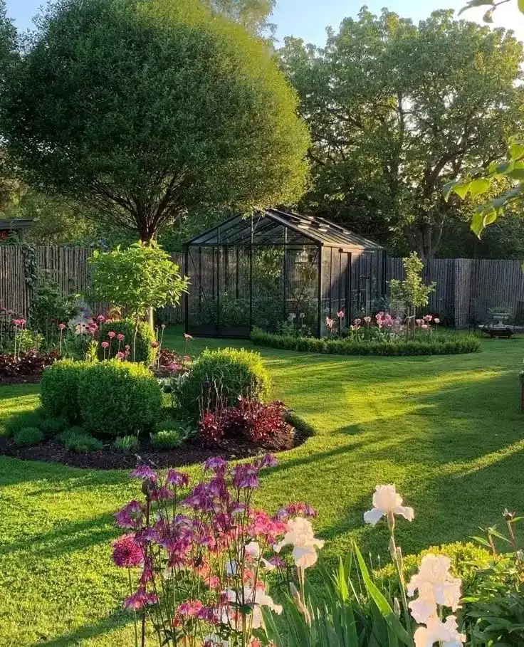Lush garden with vibrant flowers and a glass greenhouse under a tree, bathed in warm sunlight.