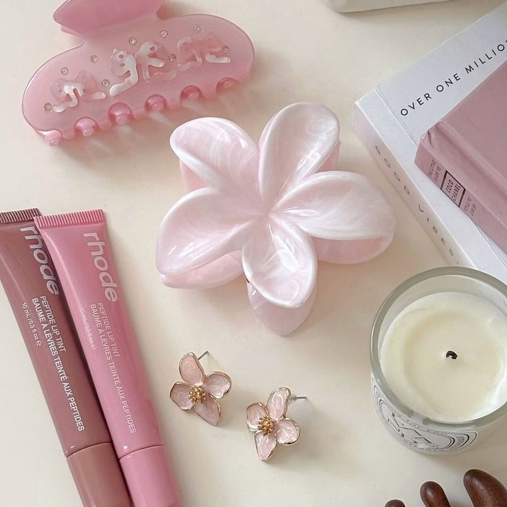 Pink beauty accessories with hair clips, lip tint, earrings, books, and a candle arranged aesthetically on a light background.