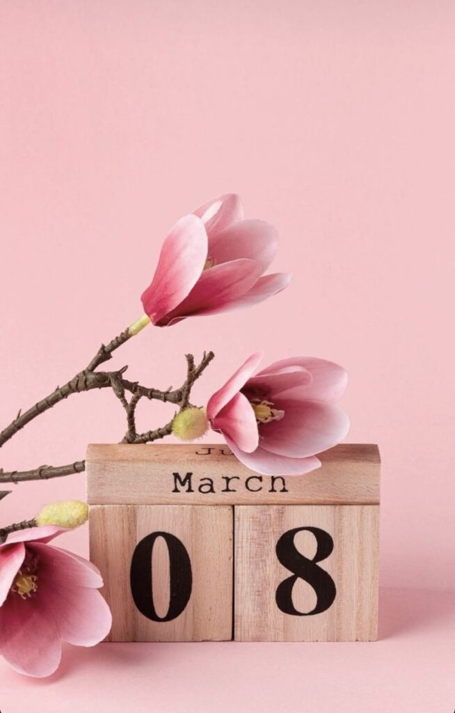 Pink magnolias and wooden calendar displaying March 8 on pastel pink background, celebrating International Women's Day.