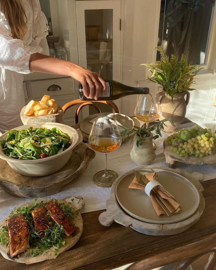 Elegant dining setup with fresh salad, grilled salmon, baguette slices, and wine. Cozy sunlight ambiance and decorative greenery.