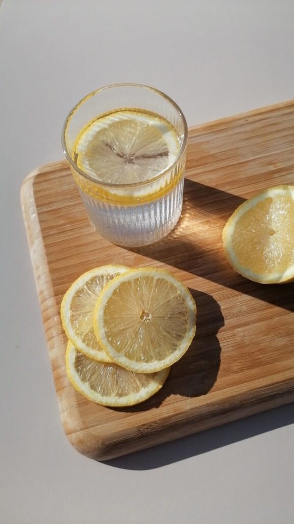 Glass of lemon water on wooden board with fresh lemon slices, sunlight casting shadows. Refreshing and healthy drink concept.