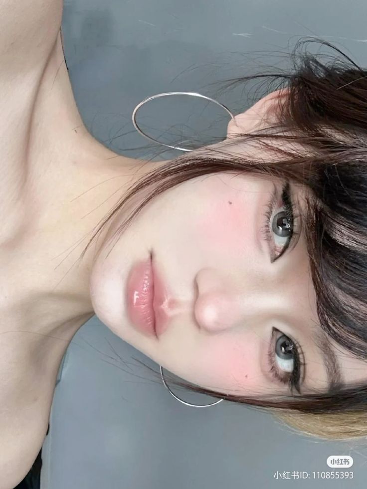 Close-up of a young person with hoop earrings, soft makeup, and brown hair against a gray background. Elegant and serene expression.