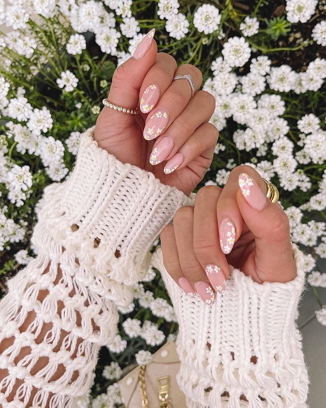 Floral nail art with pink polish and sequins, paired with a white crocheted sweater, surrounded by blooming white flowers.