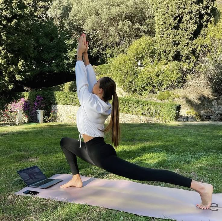 Woman practicing yoga in a lunge pose on a mat outdoors, with a laptop nearby for a virtual class session.