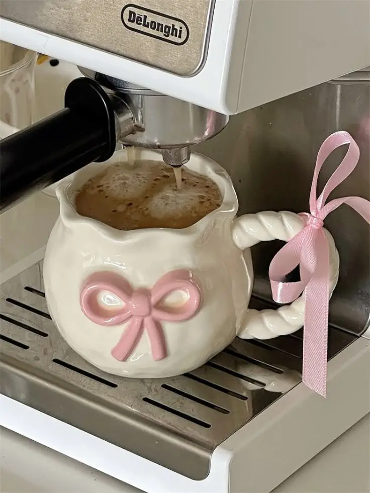 Espresso pouring into a cute ceramic mug with a pink bow from a DeLonghi machine. Perfect morning coffee setup.