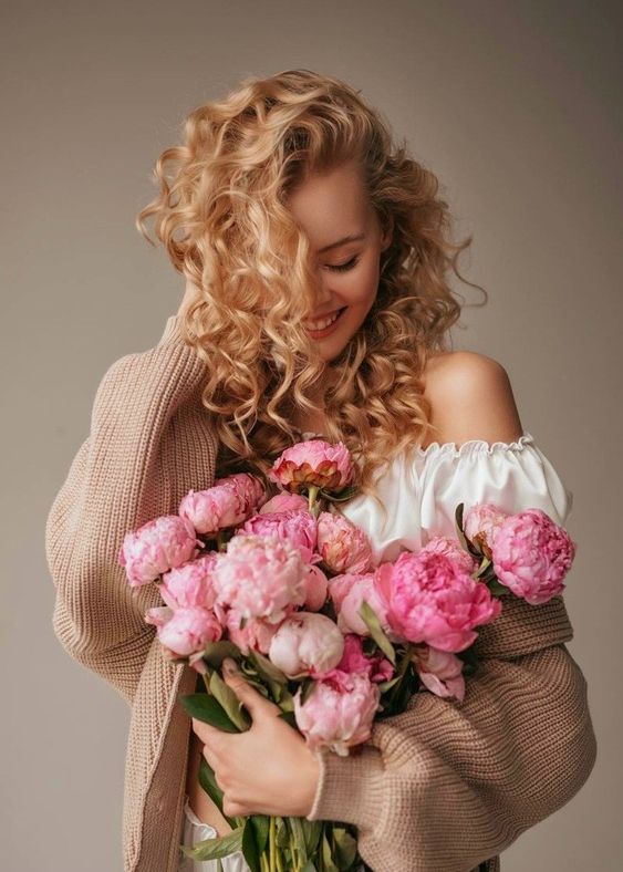 Woman with curly hair smiling, holding a bouquet of pink peonies, wearing a cozy sweater in a soft, elegant setting.