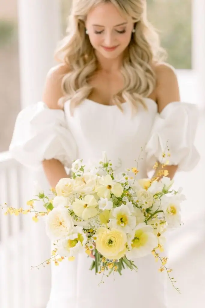 Bride in off-shoulder white dress holding bouquet of yellow and white flowers, smiling gently. Perfect for wedding inspiration.