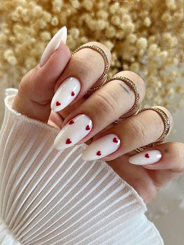 Stylish hand with white nails featuring tiny red hearts, adorned with delicate gold rings, against a textured background.