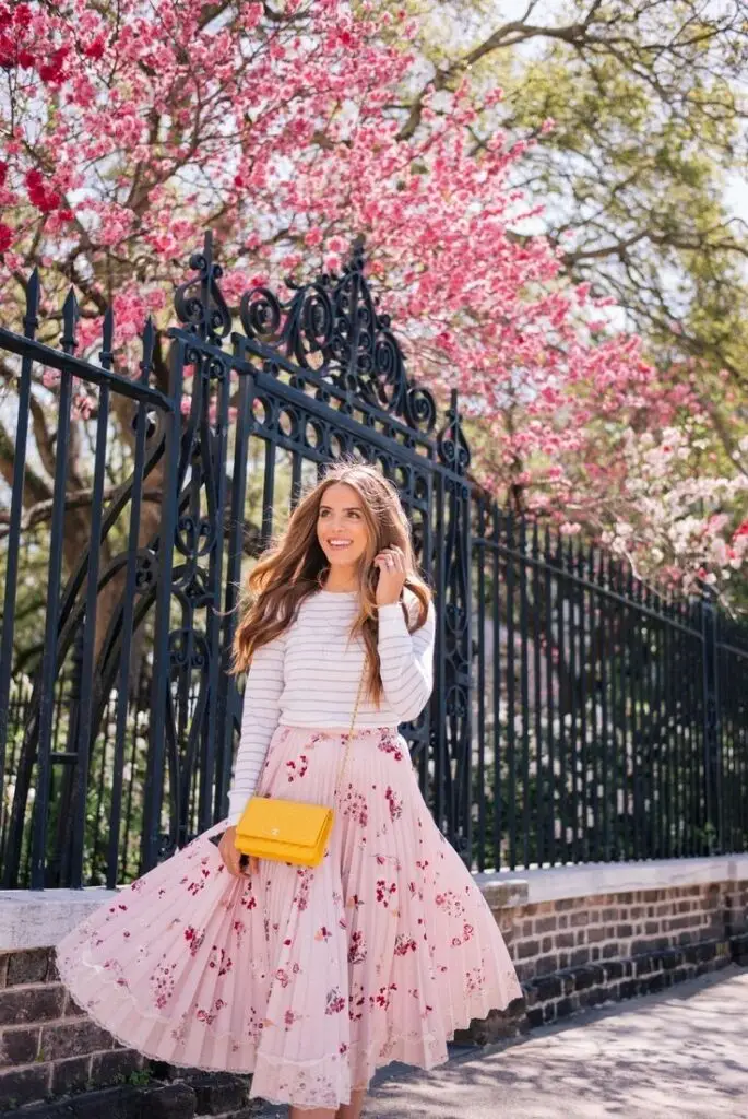 Woman in floral skirt with yellow purse walks by pink blossoming trees and ornate fence, enjoying a sunny spring day.