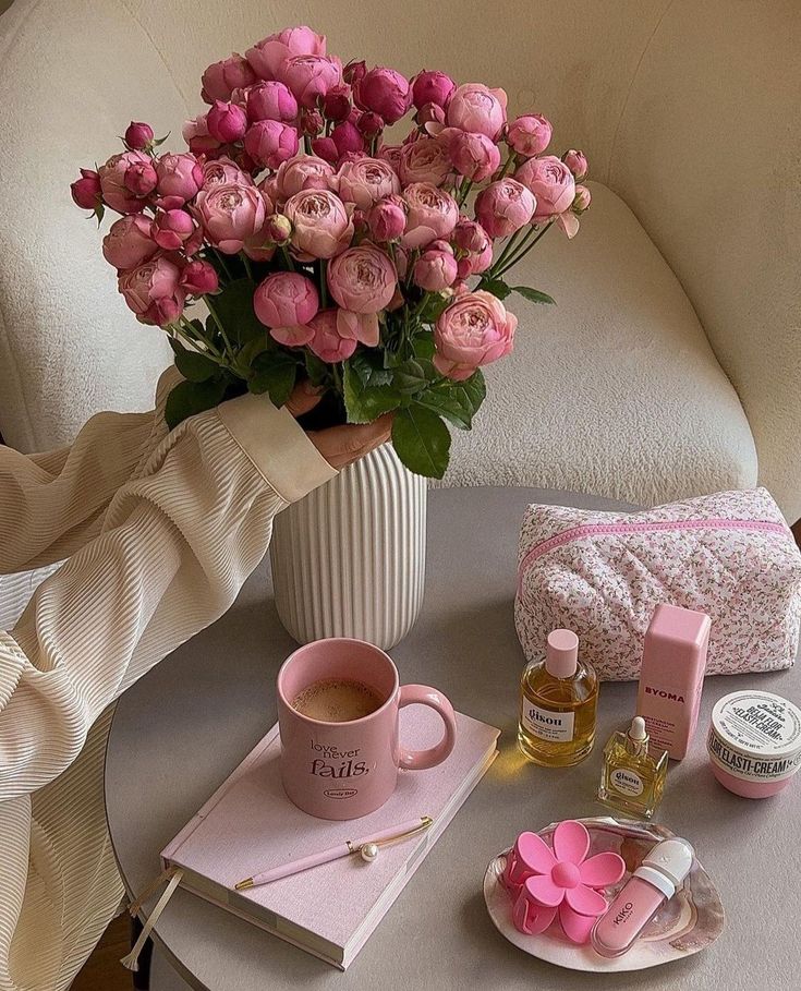 Pink roses in a vase beside a mug, notebook, cosmetics, and a floral pouch on a cozy table setting. Elegant and relaxing atmosphere.